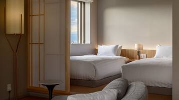 A hotel room with two beds, a standing lamp, a small table, and a view of the city through a window.