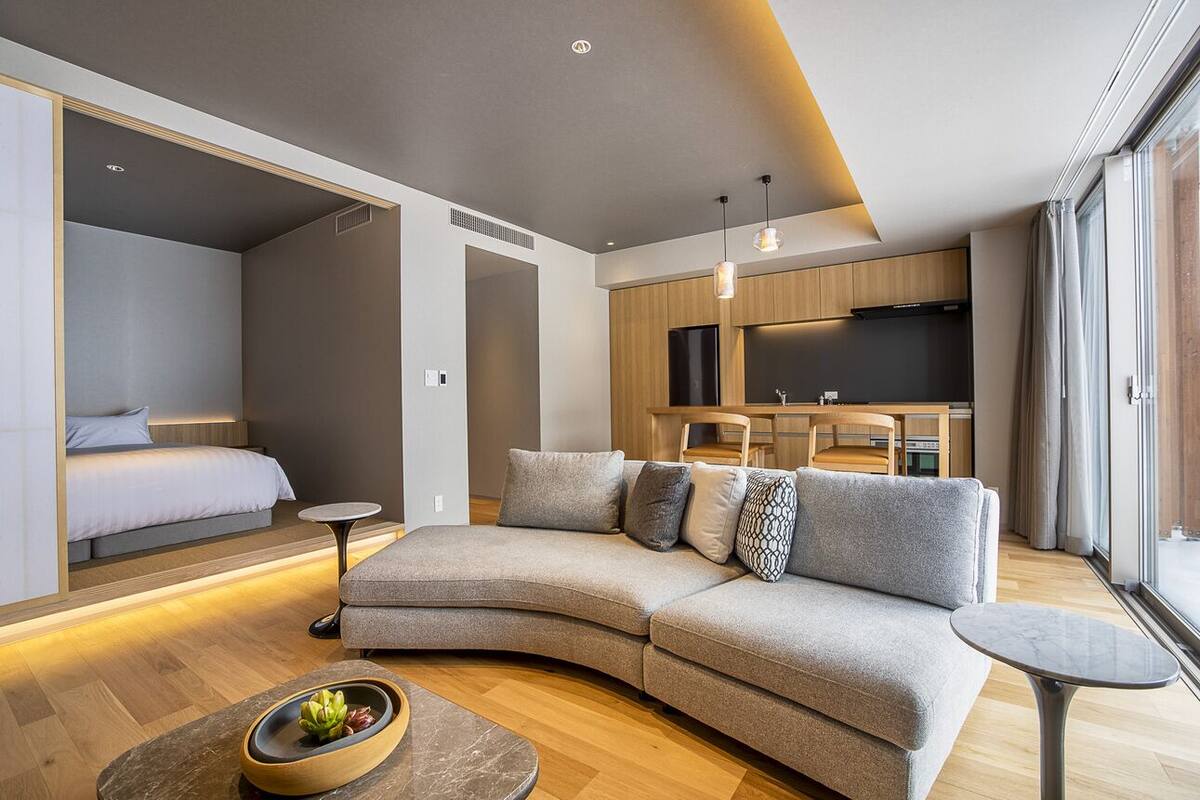 A modern living room with a sectional sofa, a small round table, and a kitchen area with wooden cabinets and a TV.
