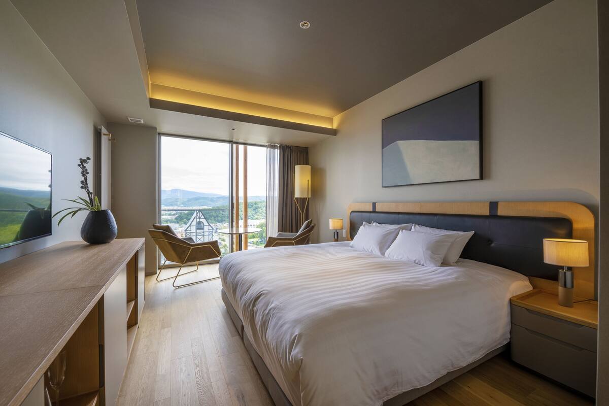 A modern hotel room with a large bed, a desk, a chair, and a view of the city.