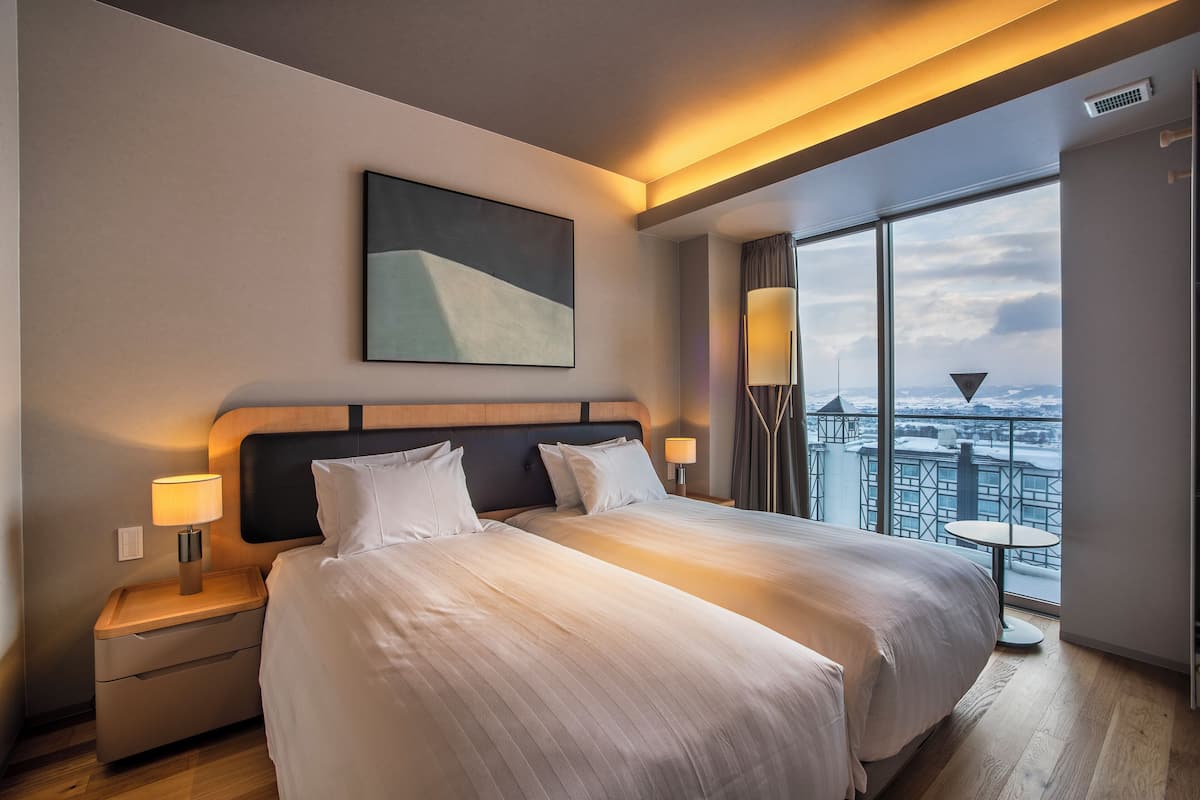 A hotel room with a large bed, two bedside tables with lamps, a view of a cityscape through large windows, and a modern abstract painting on the wall.