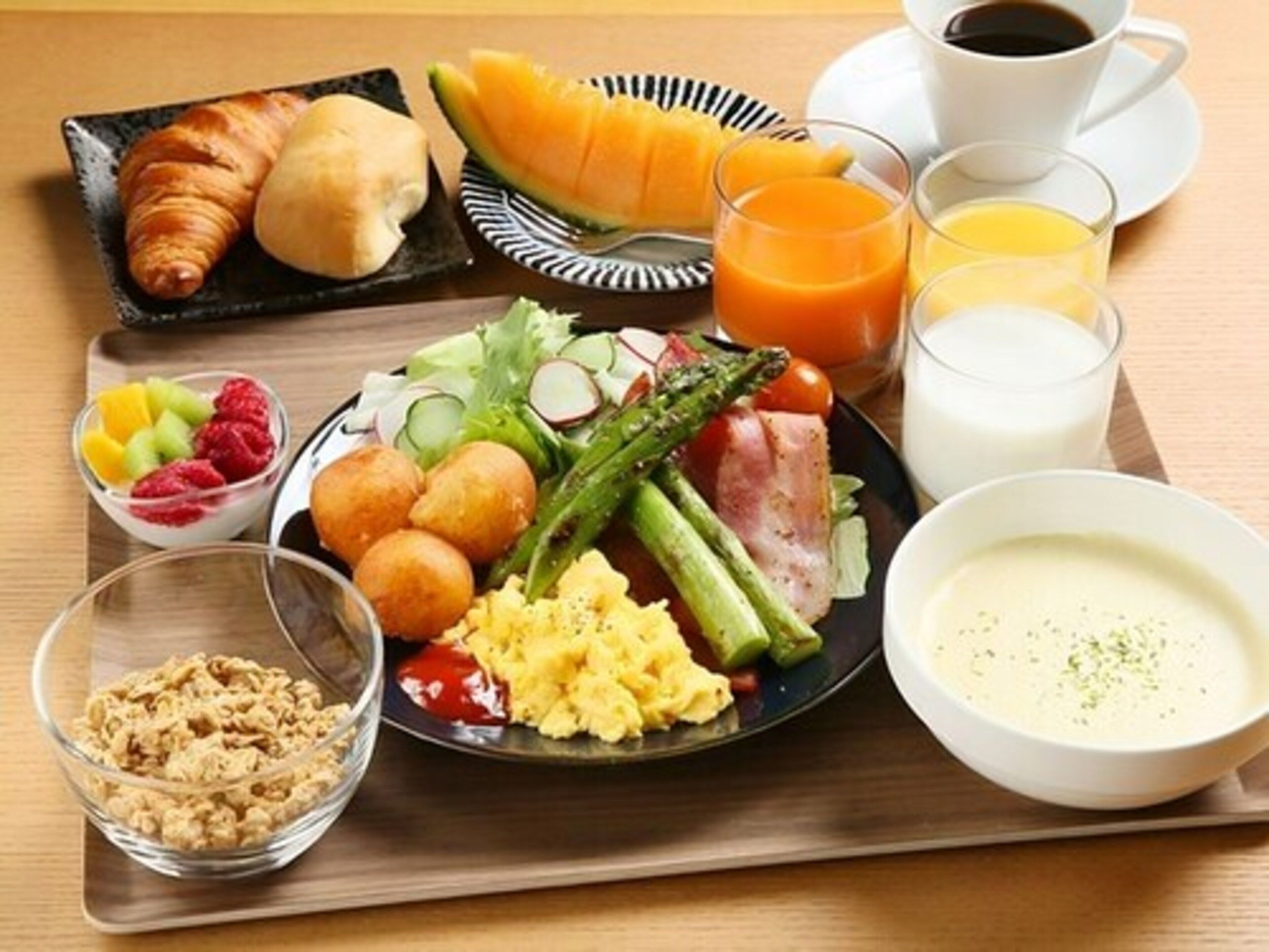 A breakfast tray with croissants, melon, scrambled eggs, asparagus, ham, and a bowl of soup.