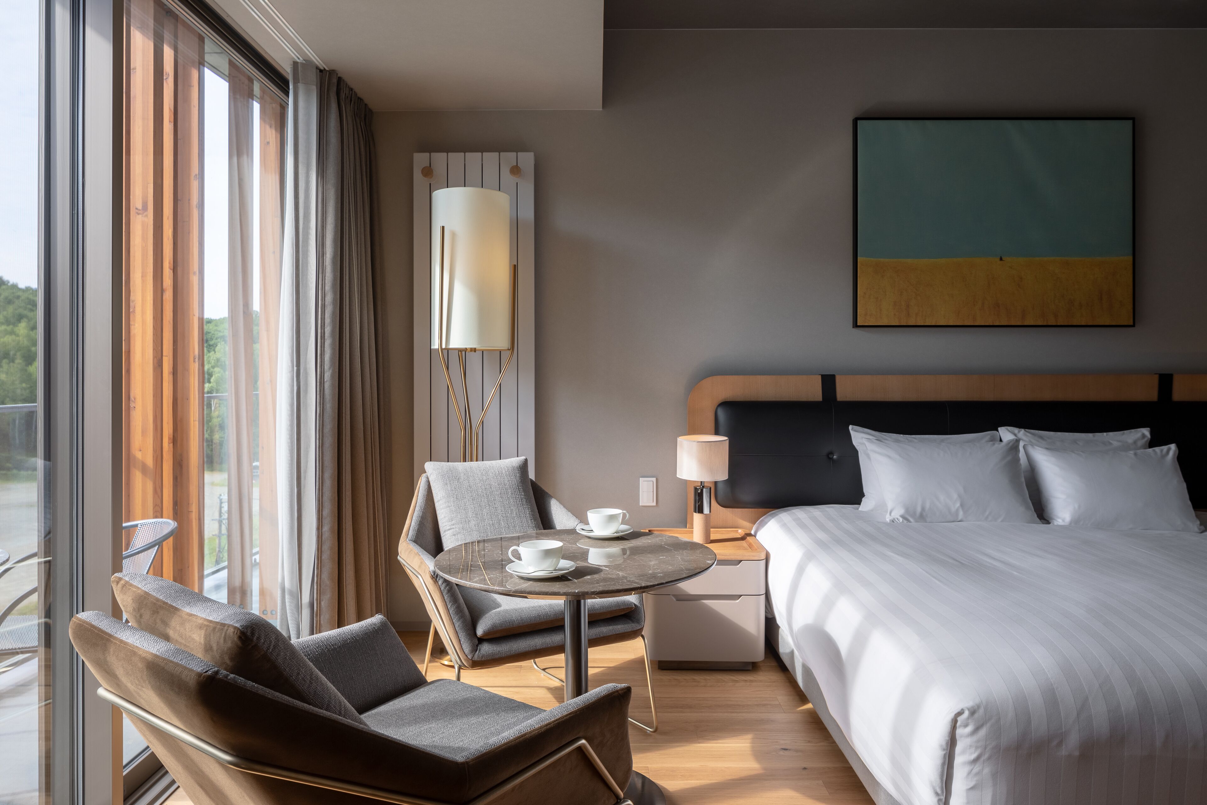 A hotel room with a bed, a chair, a table with cups, a lamp, and a painting on the wall.