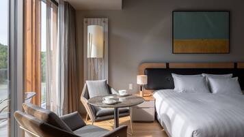 A hotel room with a bed, a chair, a table with cups, a lamp, and a painting on the wall.