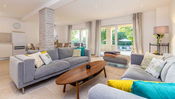 A spacious living room with a grey sofa, a wooden coffee table, and a grey armchair. There is a kitchen area with a fridge and a dining table in the background. - Holiday house VESNA with pool (Milovčići)