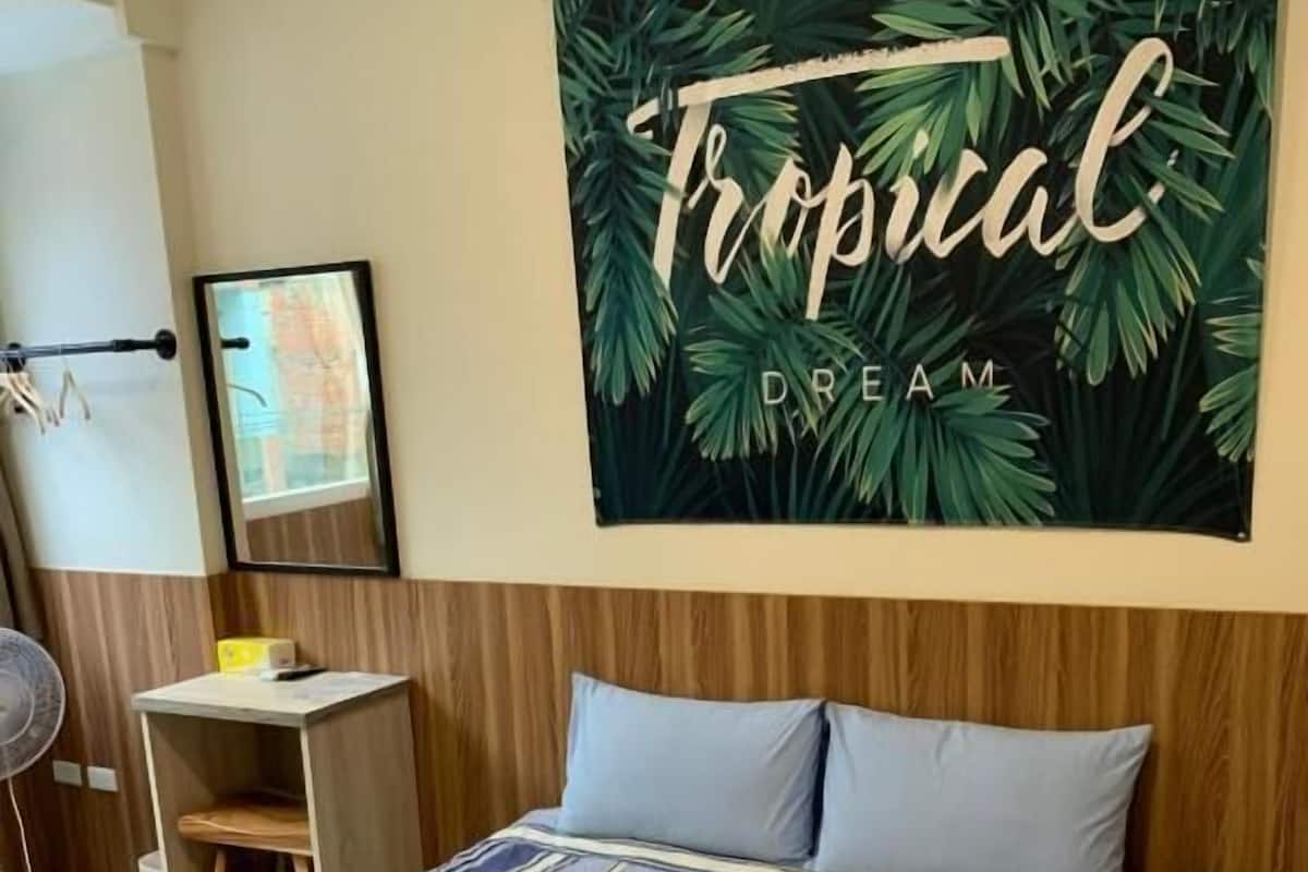 A hotel room with a bed, a small desk, a chair, and a large tropical-themed wall hanging.