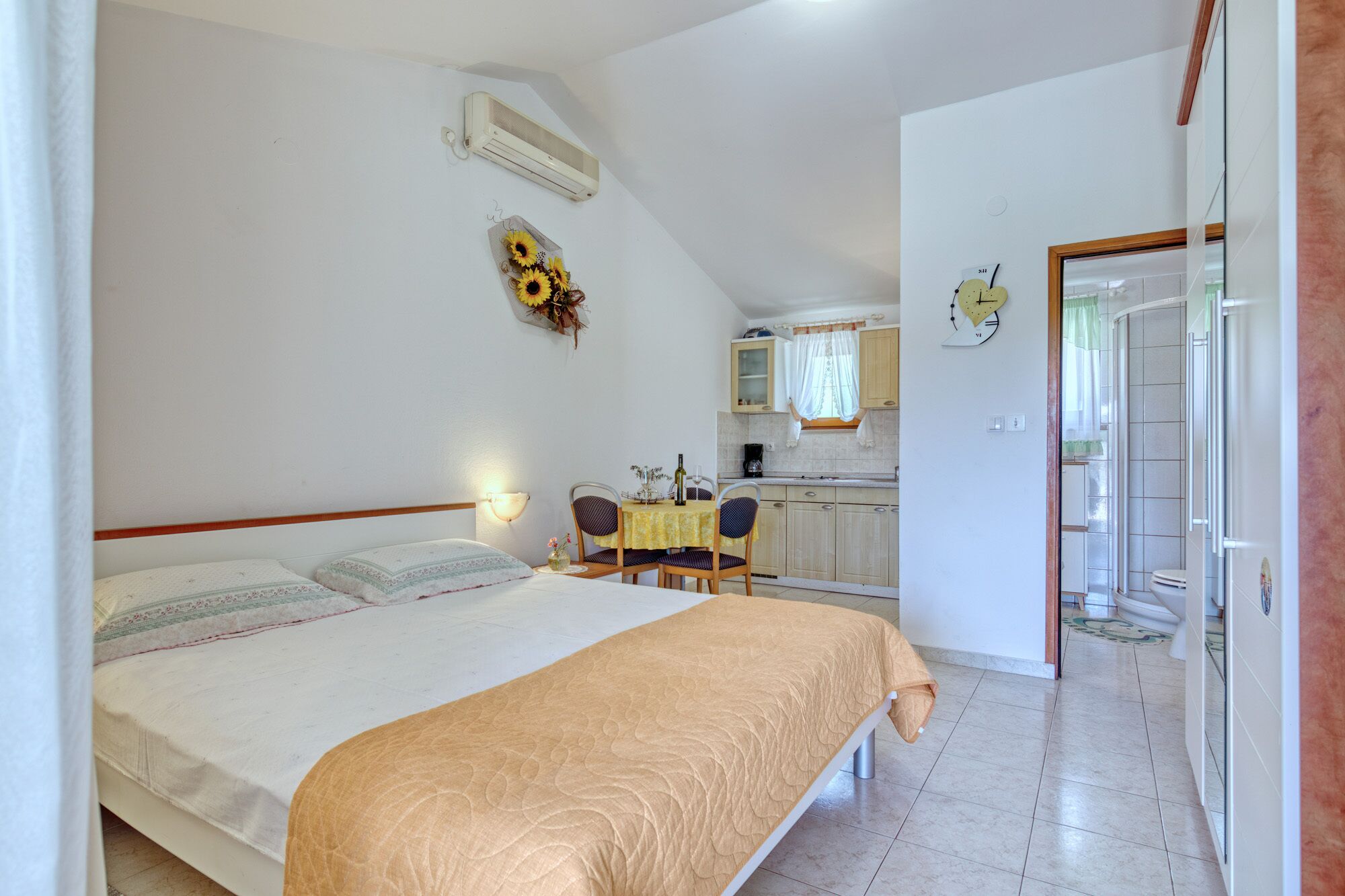 Quarto com cama, mesa pequena com cadeiras, cozinha e banheiro.