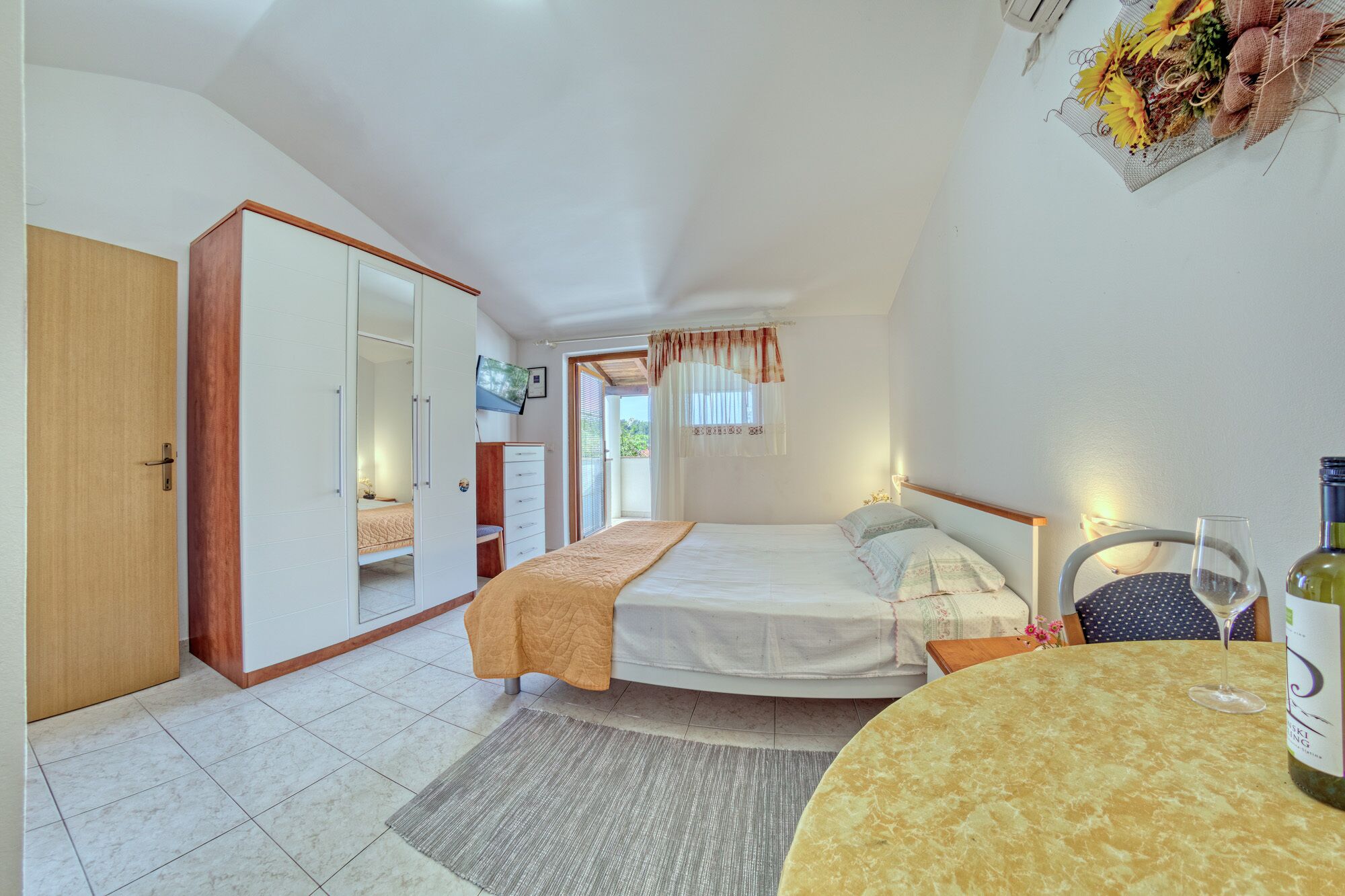 A bedroom with a bed, a desk, a chair, a table with a bottle, and a TV on the wall.