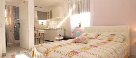 A bedroom with a large bed, a small table, and a kitchenette in the background.