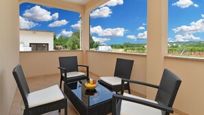 Un balcon agrémenté de meubles en osier et d’une table, un panier de fruits, et une vue sur un vignoble et des bâtiments, le tout sous un ciel bleu parsemé de nuages.