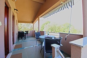 A covered balcony with a table and chairs, a door, and a view of trees.
