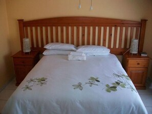 A neatly made bed with a floral bedspread, wooden headboard, two bedside tables with lamps, and a small drawer. - Koiketla Guesthouse (Akasia)