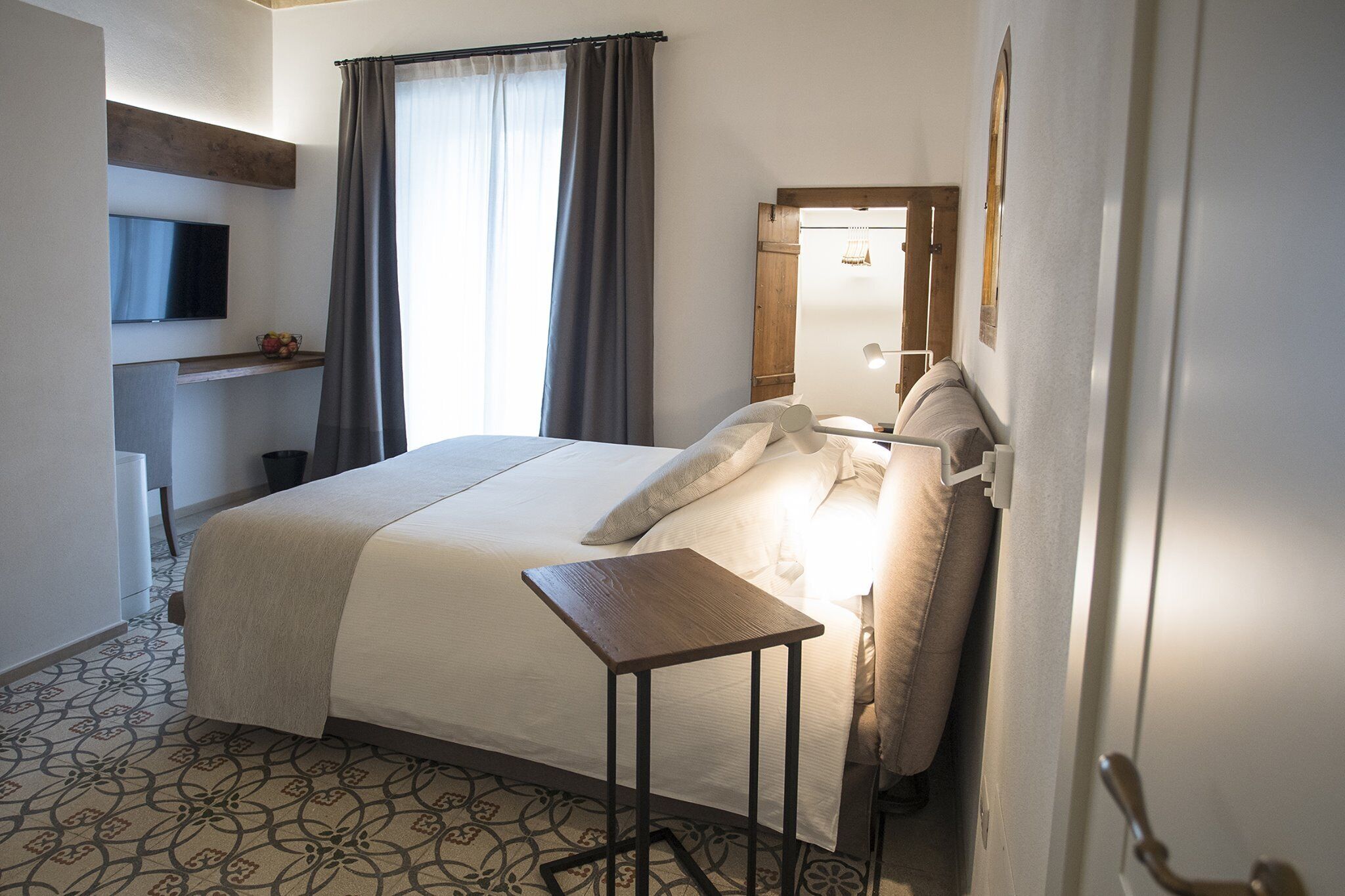 A hotel room with a bed, a bedside table, a TV mounted on the wall, and a window with curtains.