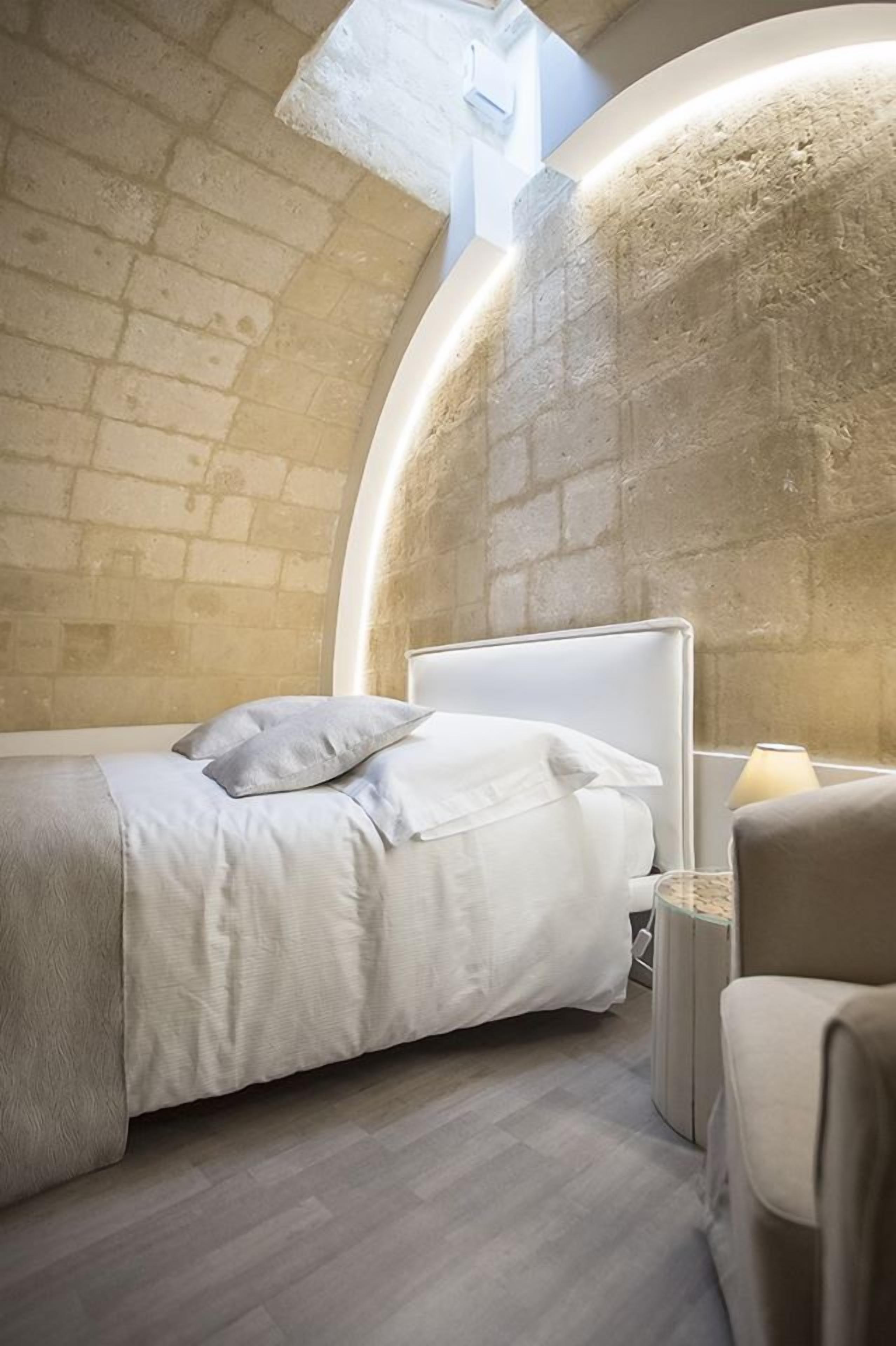 A bedroom with a curved stone wall, a bed with white bedding, a bedside table with a lamp, and a chair.