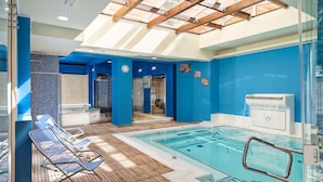 Une grande piscine intérieure dotée d’une terrasse en bois et de chaises longues, entourée de murs bleus et surmontée d’une verrière.
