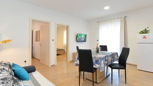 A modern living room with a sofa, a dining table with chairs, a TV mounted on the wall, and a refrigerator. - Apartment 1701/17572 (Istria - Fažana), 300m from the beach (Fažana)