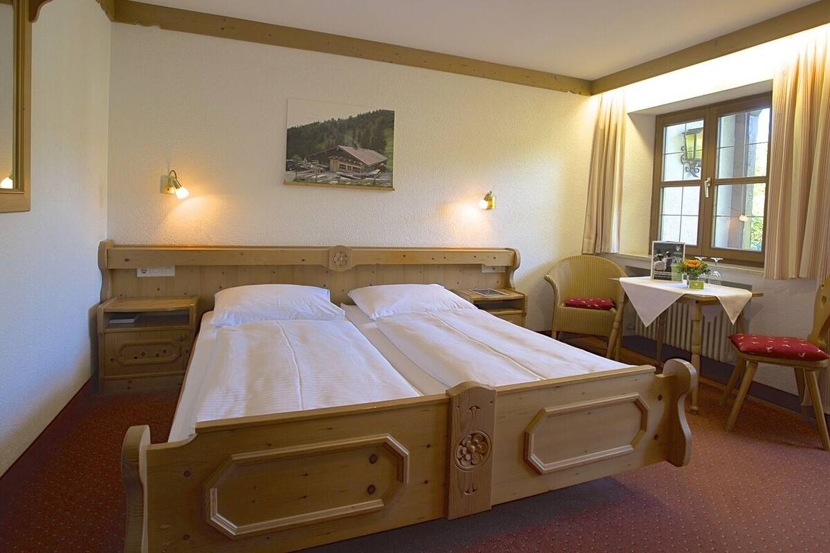 A double bed with wooden headboard, a small table with a chair, a window with curtains, and a wall-mounted picture.