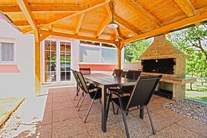 Eine Terrasse im Freien mit einem Holztisch und Stühlen, einem Grill aus Ziegelsteinen sowie einer Glastür, die zu einem Raum führt.