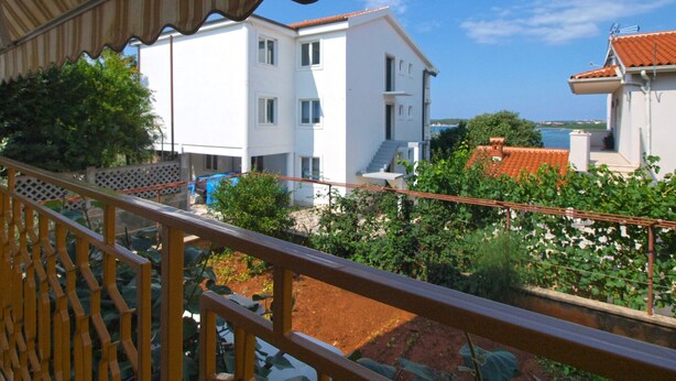 A balcony with a view of a white building with multiple windows and balconies, surrounded by greenery and a clear blue sky.