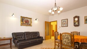 A living room with a dark brown leather sofa, a wooden dining table with floral chairs, a chandelier, and a clock on the wall.