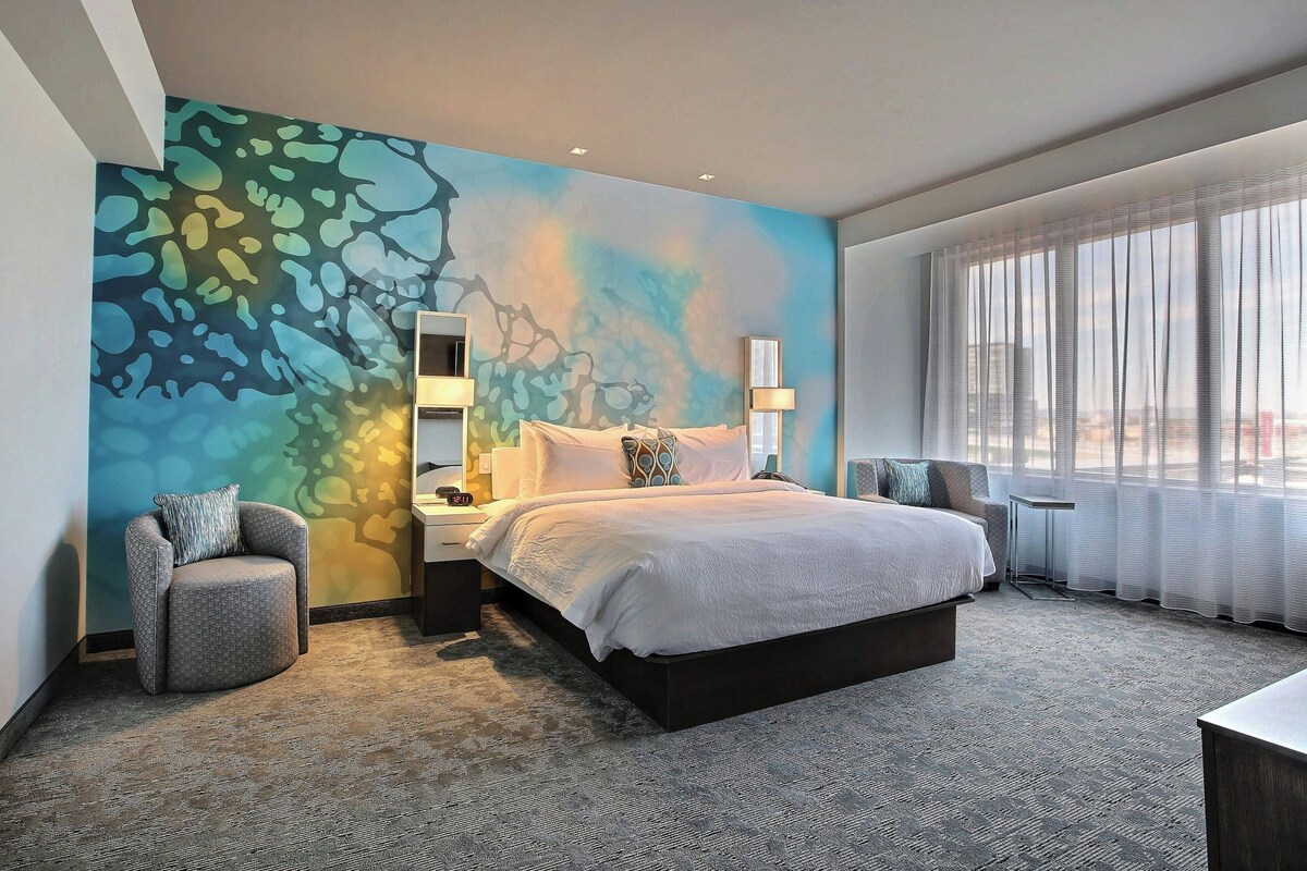Ein modernes Hotelzimmer mit einem großen Bett, einer Sitzecke, einem Wandbild und Blick auf die Stadt durch leichte Vorhänge.