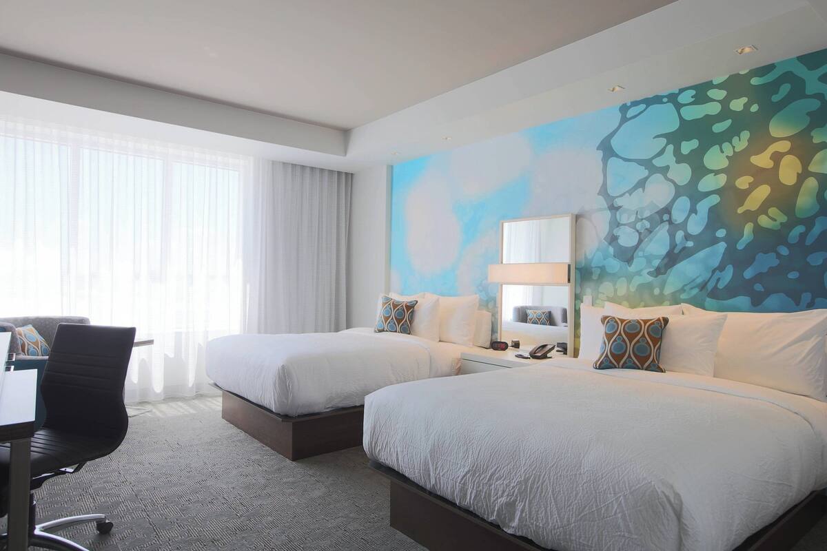 Ein Hotelzimmer mit zwei Betten, einem Schreibtisch und einem großen, abstrakten Wandgemälde.