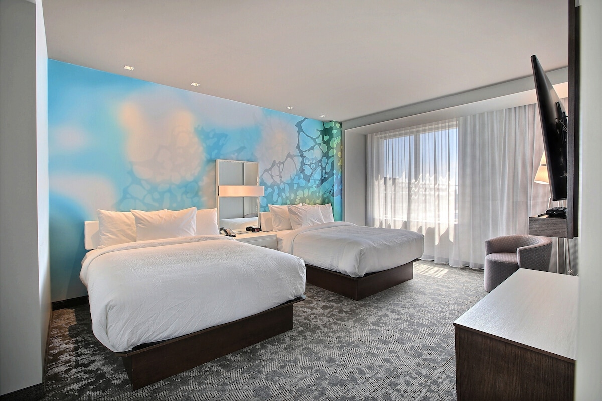 Ein Hotelzimmer mit zwei Betten, einem Schreibtisch, einem Sessel, einem Fernseher und einem großen Fenster mit Vorhängen.