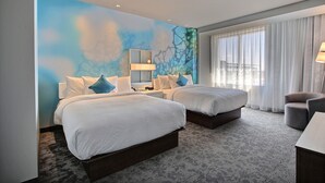 Une chambre d’hôtel avec deux lits, une grande fenêtre avec des rideaux légers, une fresque murale et un petit coin salon.