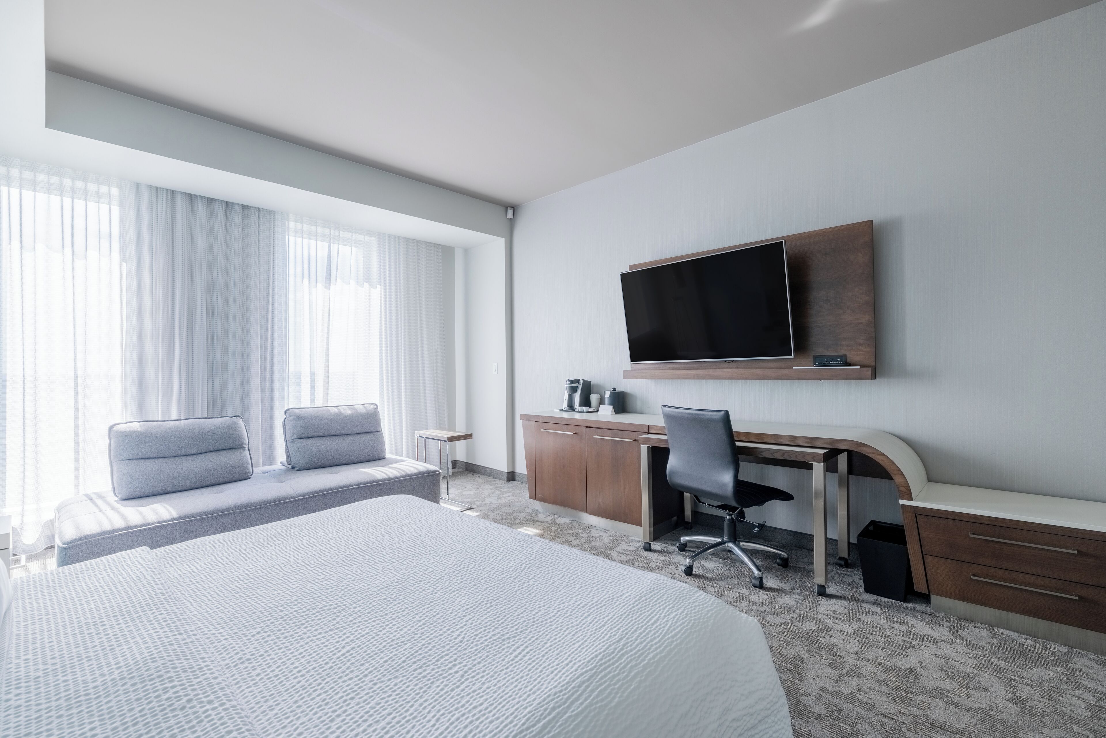Habitación de hotel con una cama grande, un escritorio con una silla, un televisor montado en la pared y una ventana con cortinas traslúcidas.