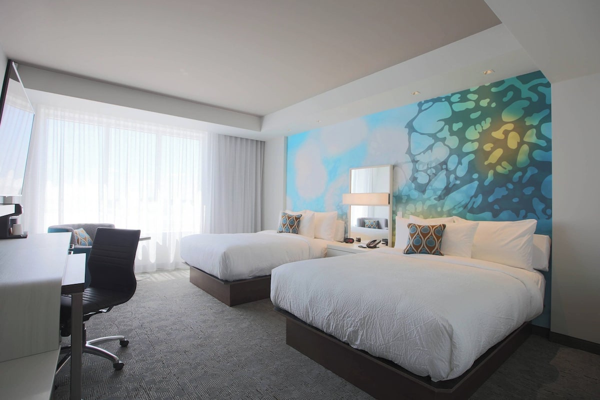 Ein Hotelzimmer mit zwei Betten, einem Schreibtisch und einem Stuhl. Es gibt ein großes Fenster mit Vorhängen, ein Wandbild und einen Nachttisch mit einer Lampe.