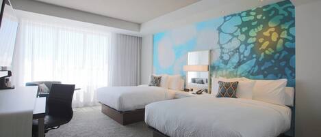 A hotel room with two beds, a desk, and a chair. There is a large window with curtains, a wall mural, and a nightstand with a lamp.