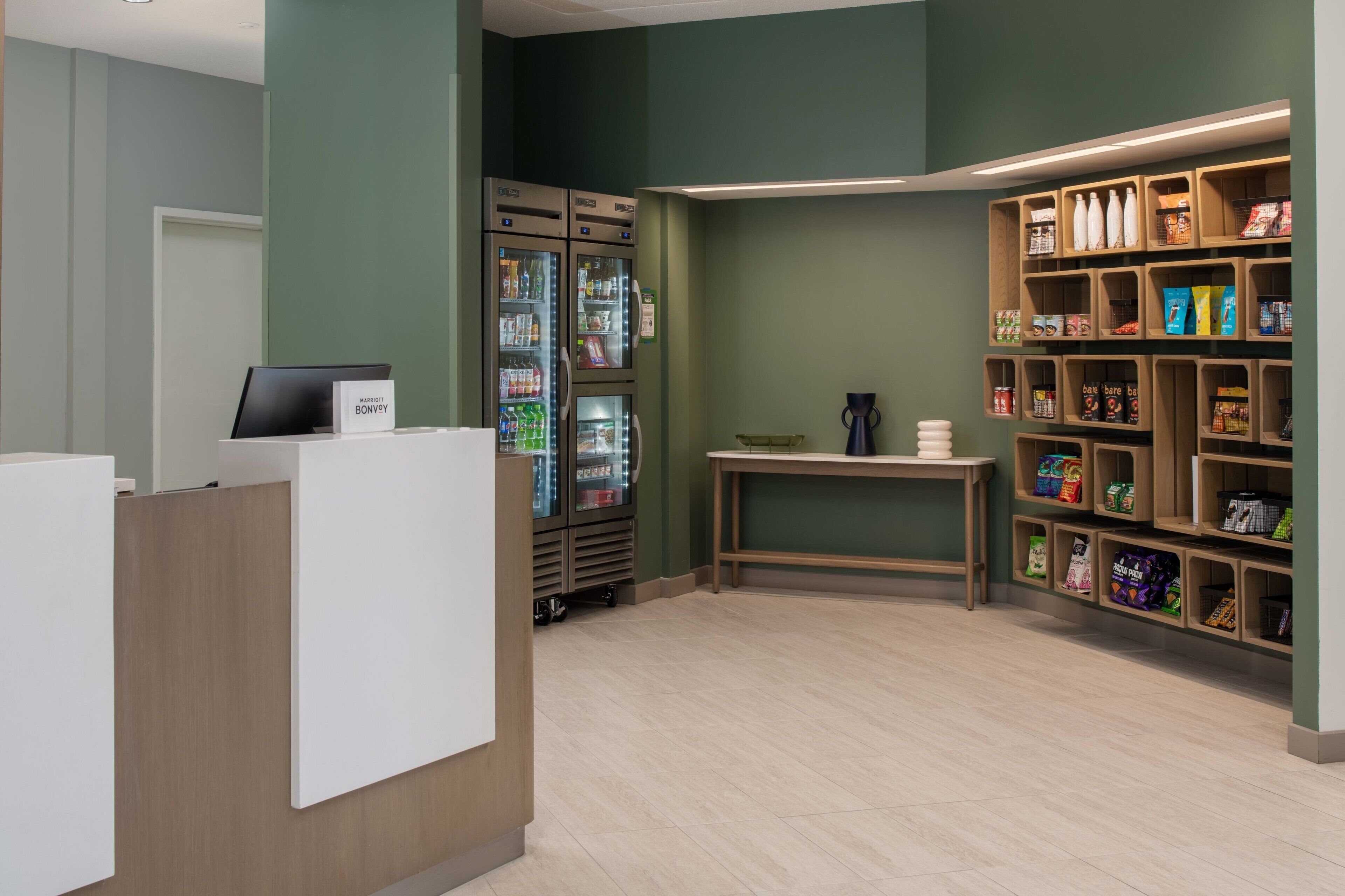 Zona de recepción de un hotel con mostrador de recepción, minibar y estante con aperitivos y bebidas.