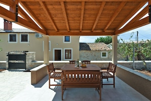 An outdoor patio with a wooden table and chairs, a stone wall, and a building with a tiled roof in the background.