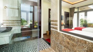 A modern hotel room with a glass partition, a bed with red bedding, a marble floor, and a view of the outdoors.