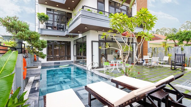 A modern two-story building with a swimming pool, outdoor seating, and lush greenery.