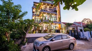 A multi-story building with balconies, a car parked in front, and surrounding greenery.