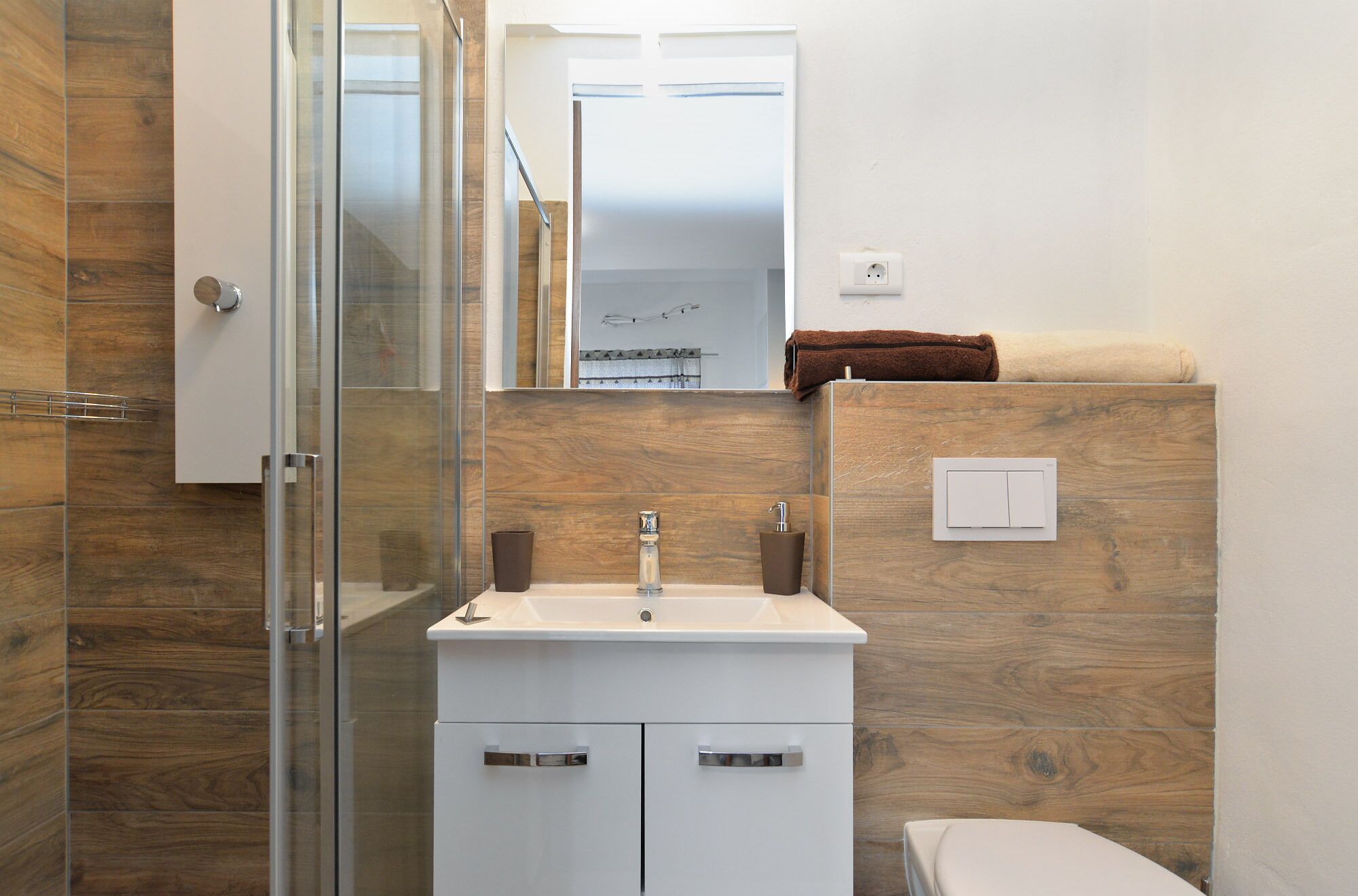 A bathroom with a glass shower enclosure, a white sink with two towel holders, and a toilet.