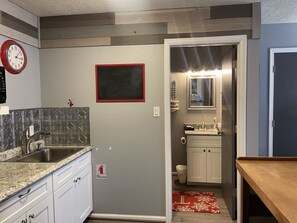 A kitchen with a granite countertop, stainless steel sink, and white cabinets. There is a red-framed mirror above the sink and a fire extinguisher sign on the wall.