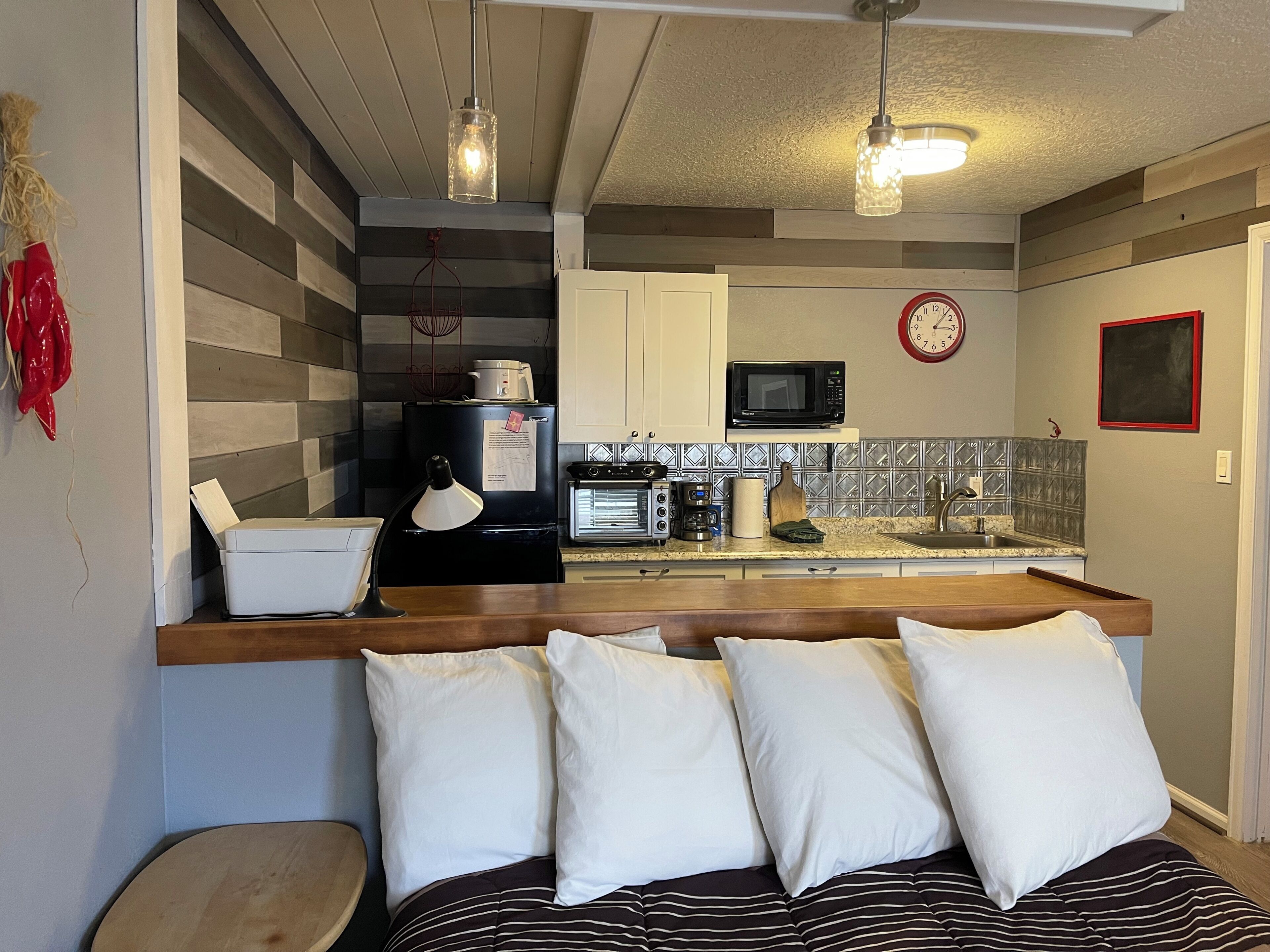 A compact kitchen with a microwave, stove, and sink. There is a clock on the wall and a red decorative item on the left.