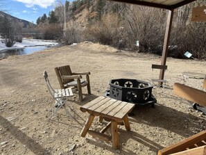 A rustic outdoor seating area with a wooden table and chairs, a fire pit, and a view of a river and mountains.
