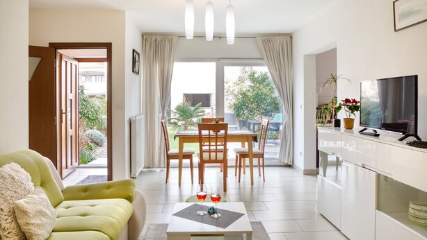 A modern living room with a green sofa, a dining table with chairs, a kitchen area with a TV, and a view of the outdoors through large windows. - House 1765/19674 (Istria - Peroj), 1000m from the beach (Peroj)