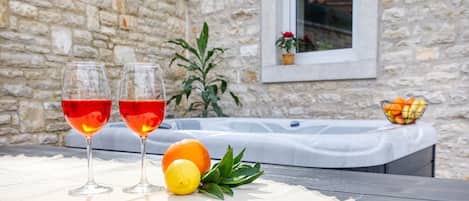 A stone wall, a hot tub, and a table with two glasses of red wine, an orange, and a lemon.