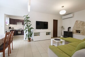Un salon moderne avec un canapé vert, une table basse blanche et un téléviseur. On y trouve également un coin cuisine avec des meubles sombres et une cuisinière. Une grande plante en pot est placée dans un coin.