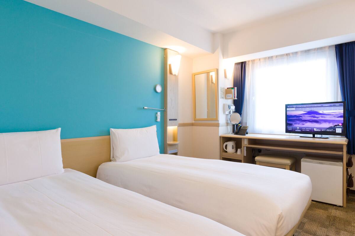 Een hotelkamer met twee bedden, een bureau met een computer, een koffiezetapparaat en een raam met gordijnen.