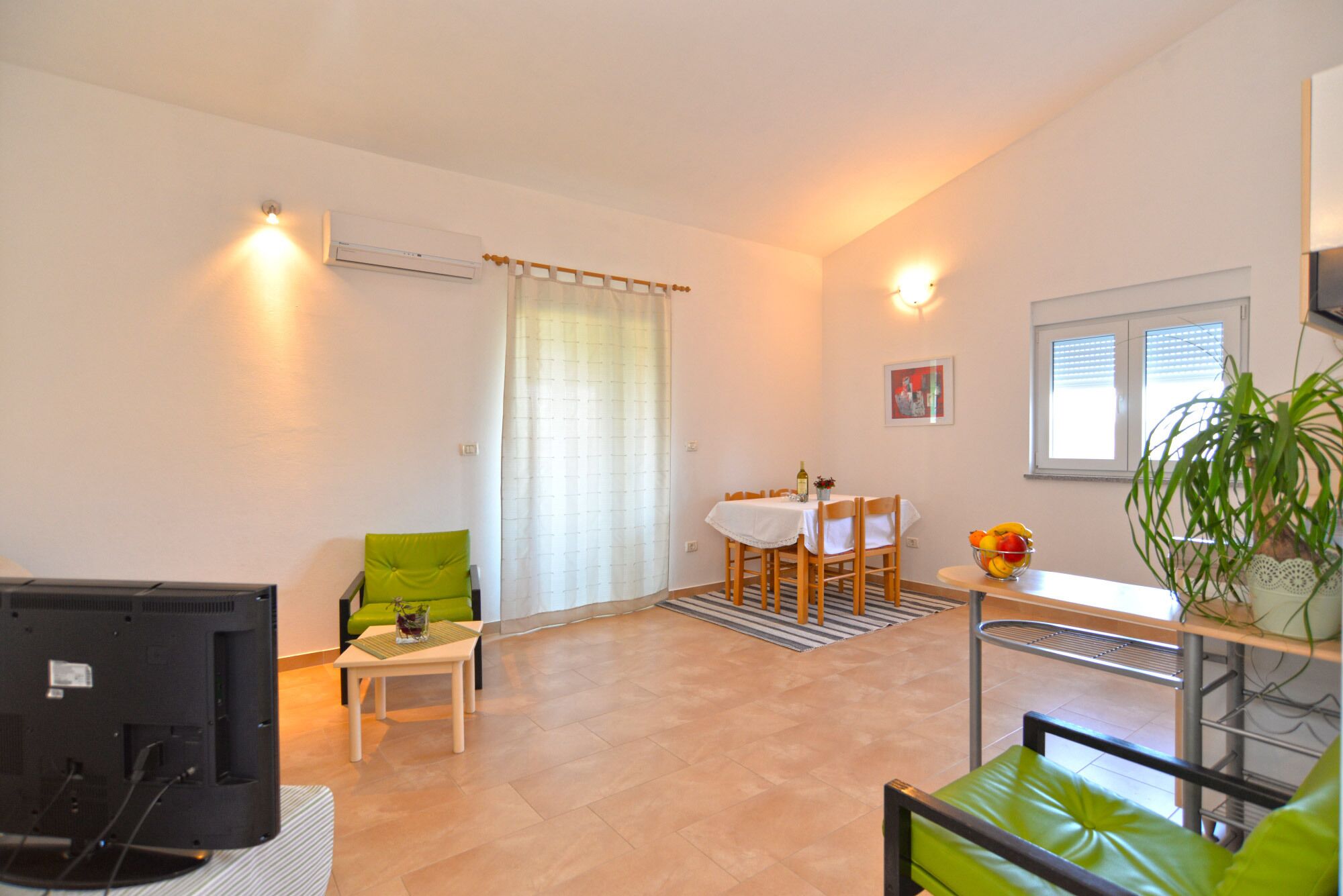 A living room with a television, a dining table with chairs, a sofa, and a potted plant.