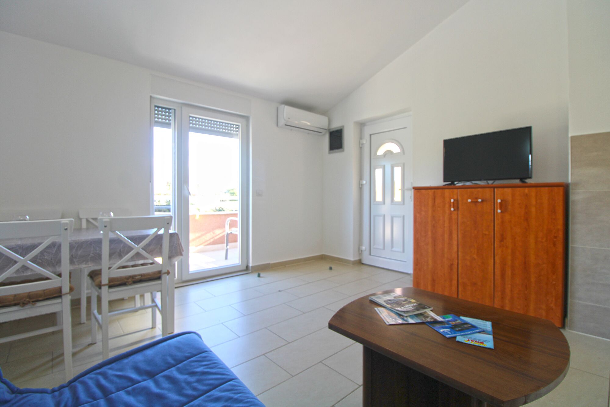 A room with a wooden table, a TV on a cabinet, a door, and a window with blinds.