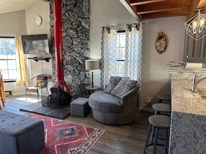 Una sala de estar con pared de piedra, chimenea, sofá, televisor y una zona de cocina con encimera de mármol.