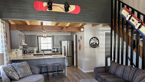 A modern kitchen with a central island, a dining area with a sofa and bar stools, and a staircase with a snowboard decoration. - Riverbend 8A Townhouse (Red River)