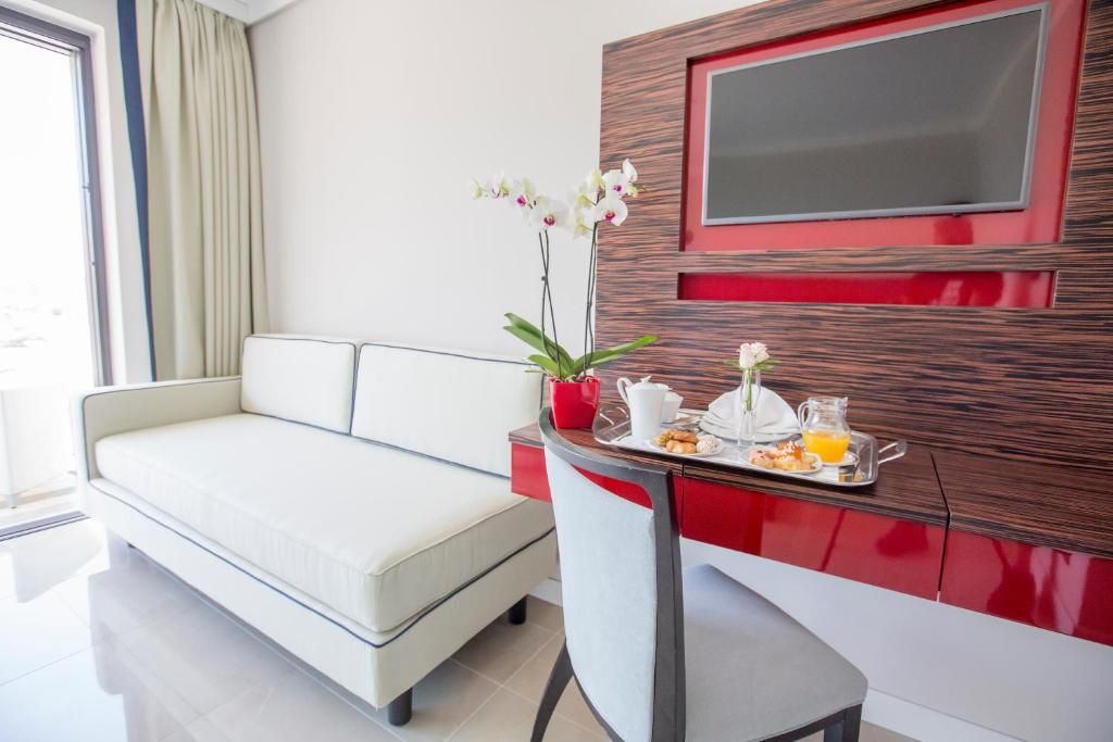 Habitación de hotel moderna con sofá blanco, mesa de comedor con alimentos para el desayuno, una orquídea en maceta y televisor montado en una pared de madera con paneles rojos.