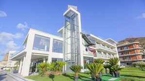 A modern building with a glass elevator shaft, large windows, and a green lawn with potted plants. - Victoria Palace Hotel (Cefalù)