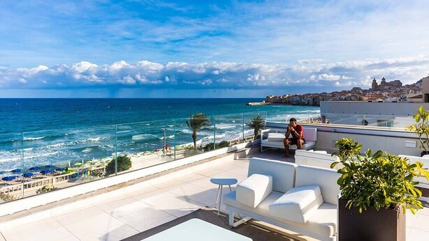 A rooftop terrace with a view of the sea, white outdoor furniture, and a potted plant. - Victoria Palace Hotel (Cefalù)